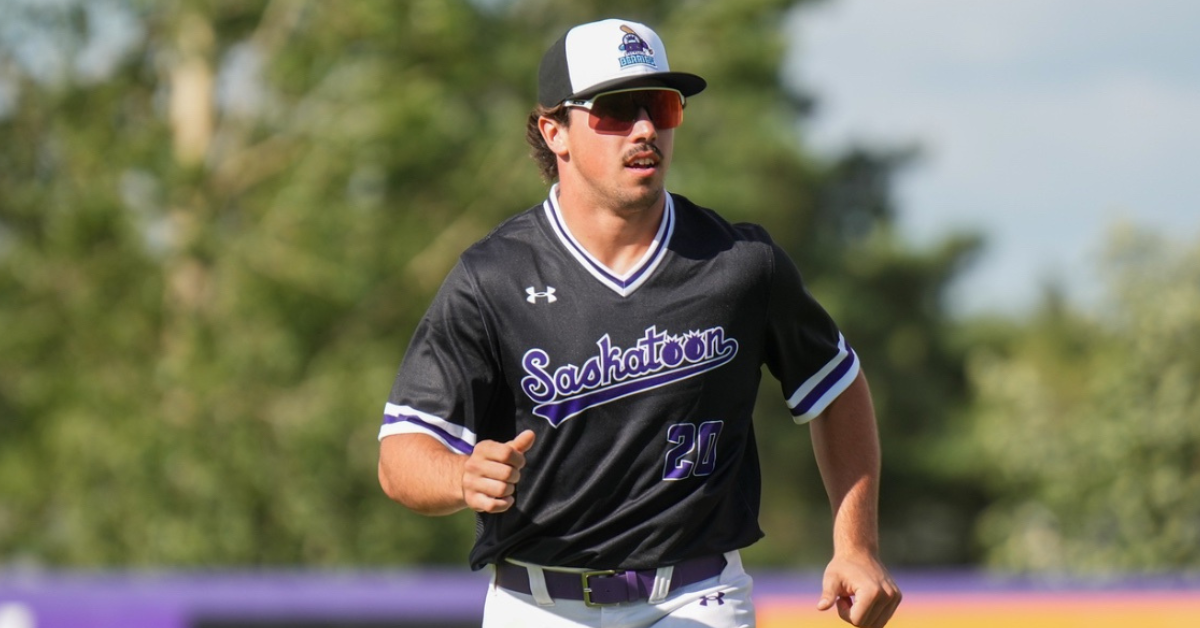 Carter beck running on baseball field. He is wearing a black Saskatoon Berries shirt, sunglasses and a white baseball cap with the Saskatoon Berries logo on it.