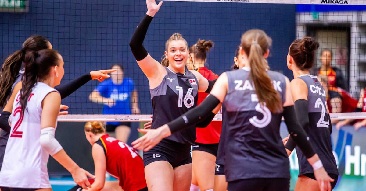 A volleyball team wearing black and grey jersey are coming together on the court. Abby Wasutyk has arm raised while facing the camera smiling to high five teammate.